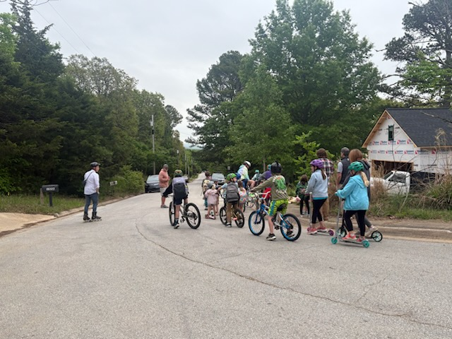 People riding bicycles and scooters on a street surrounded by trees and houses. A man stands nearby.