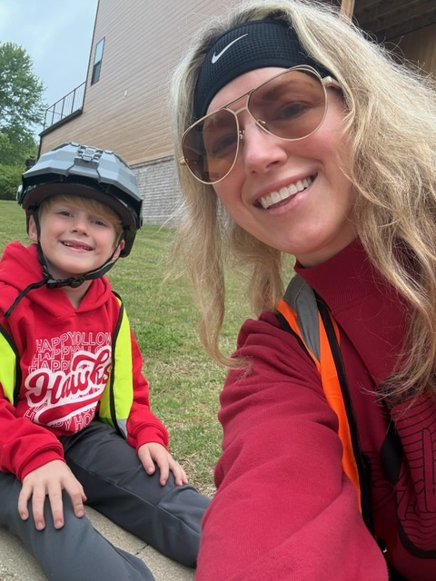 A woman in a red shirt and sunglasses poses with a smiling child wearing a helmet.
