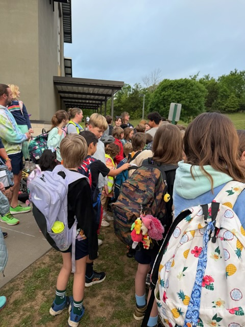 A crowd of children, wearing backpacks, standing outside near a building on a sunny day.