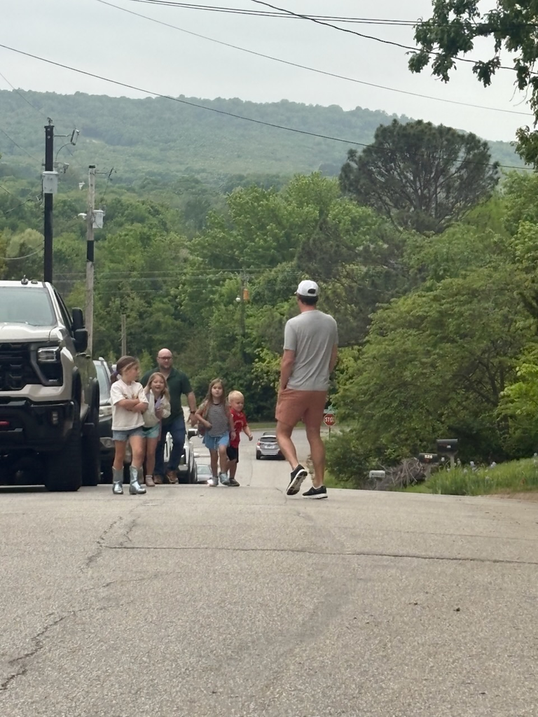 A group of people, including adults and children, walk down a road with trees and mountains in the background.