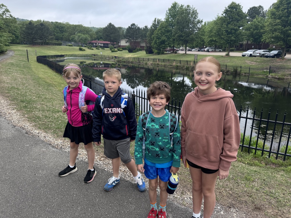Five children stand by a sidewalk. A pond with a black fence is behind them. The children wear backpacks and sneakers.