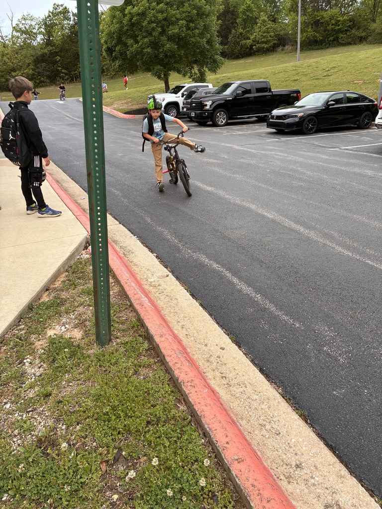 Two people standing on a sidewalk next to a road, one in a helmet riding a bicycle.