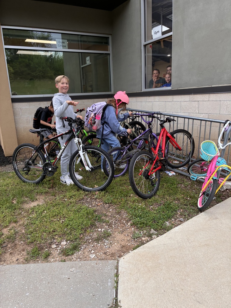 Two children with bikes near a building, one wears a pink helmet, and another has a backpack.