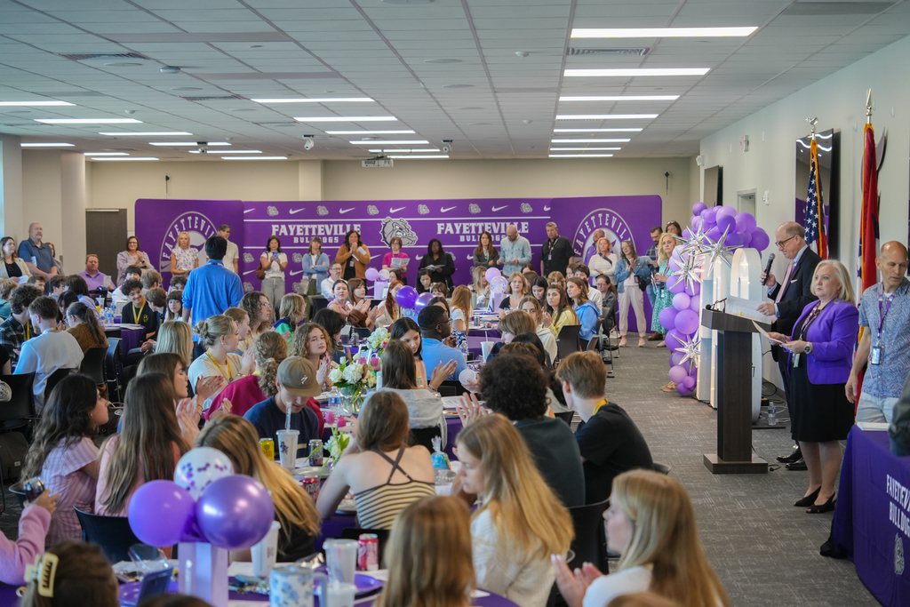 A group of people sit at tables with purple balloons and decorations in a large room.