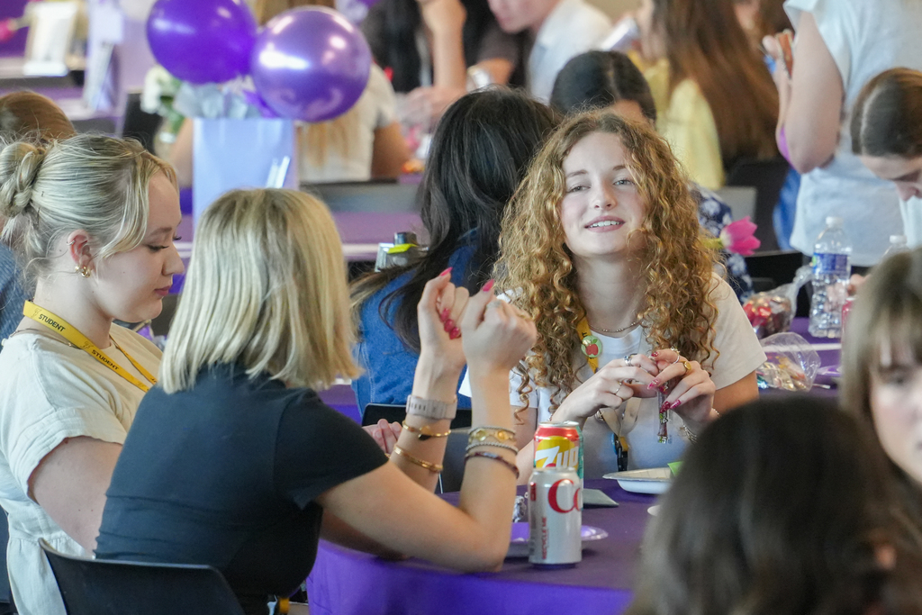 Several people seated around a table with purple tablecloth, engaged in conversation. A woman in the center smiles.