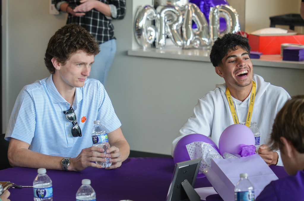 Two men seated at a table with balloons, one holding a water bottle. Another person is partially visible.