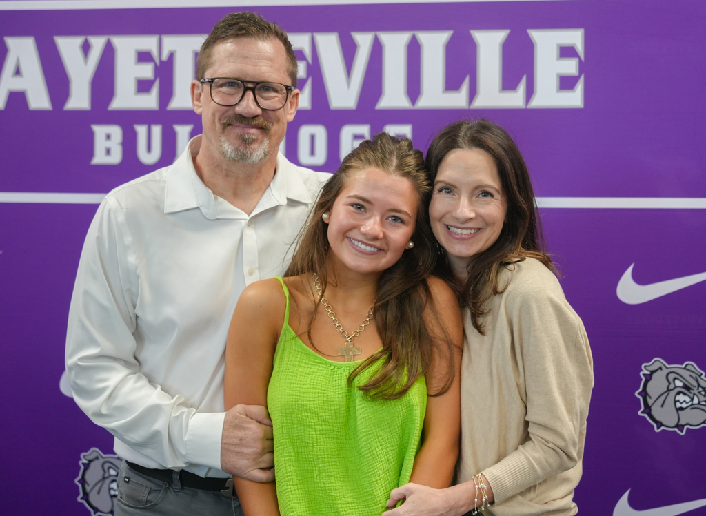 Three people stand close together, smiling, with a purple wall behind them. The wall has the words "FAYETTEVILLE BULLDOGS" and a Nike logo.