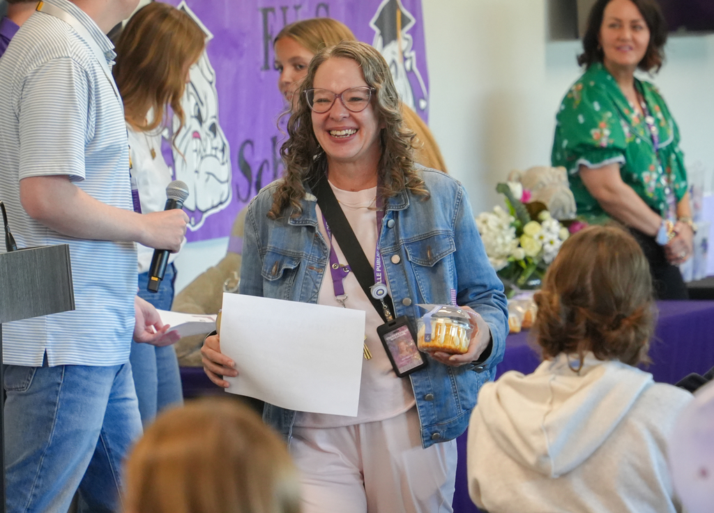Woman wearing glasses and a denim jacket holds paper and cake. People in a room with a table, flowers, and a purple banner.