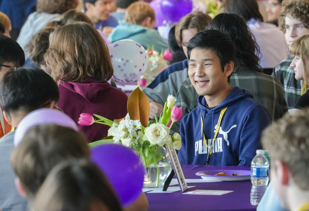 A young man in a blue hoodie smiles at a gathering. Many people sit around him, some holding purple balloons.