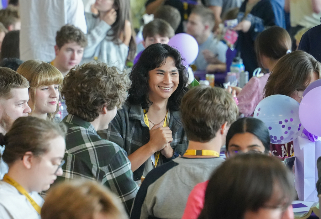 A large group of people inside a room. Some are sitting and standing. A smiling woman is surrounded by people.