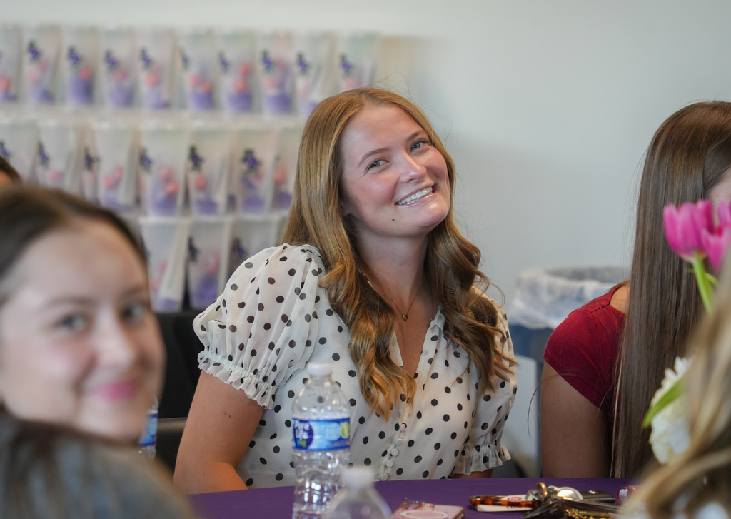 A group of women seated at a table. One woman wears a polka-dot blouse. They all smile.
