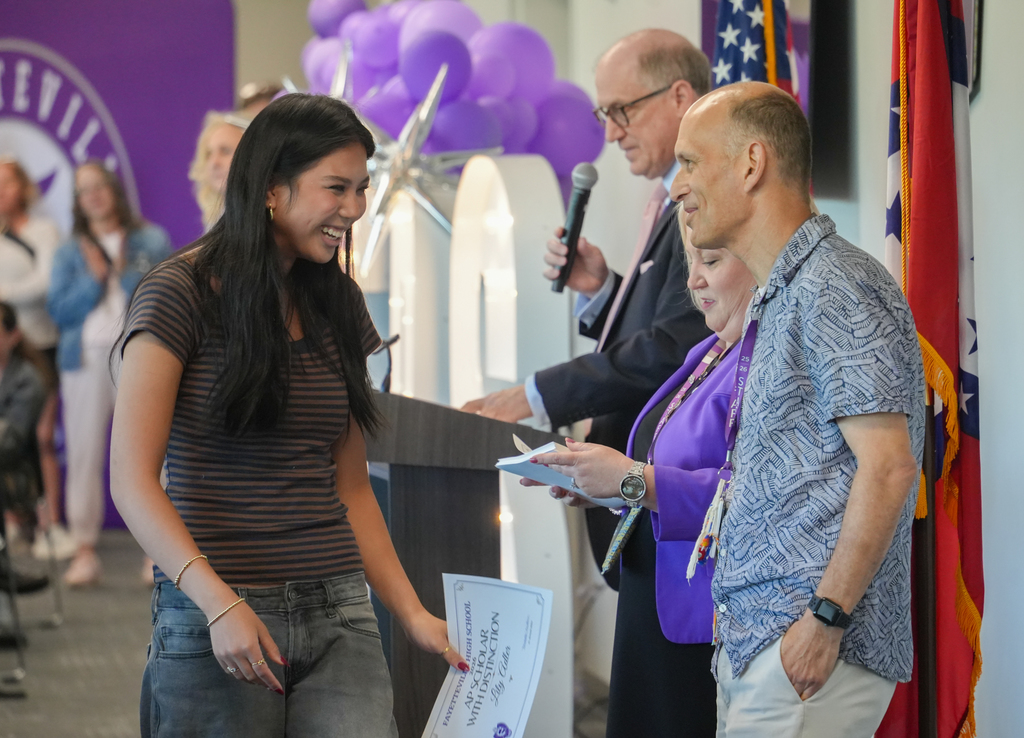 A woman in a striped shirt receives a paper award from a man with a microphone.