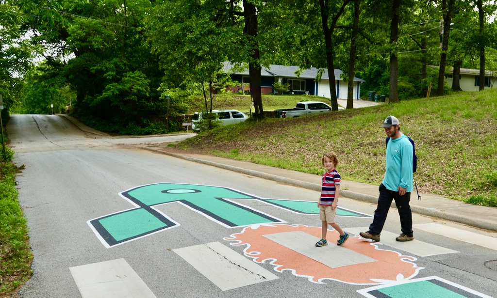 A man and a child cross a road with a large painted number 50 on it. Trees and a house are in the background.