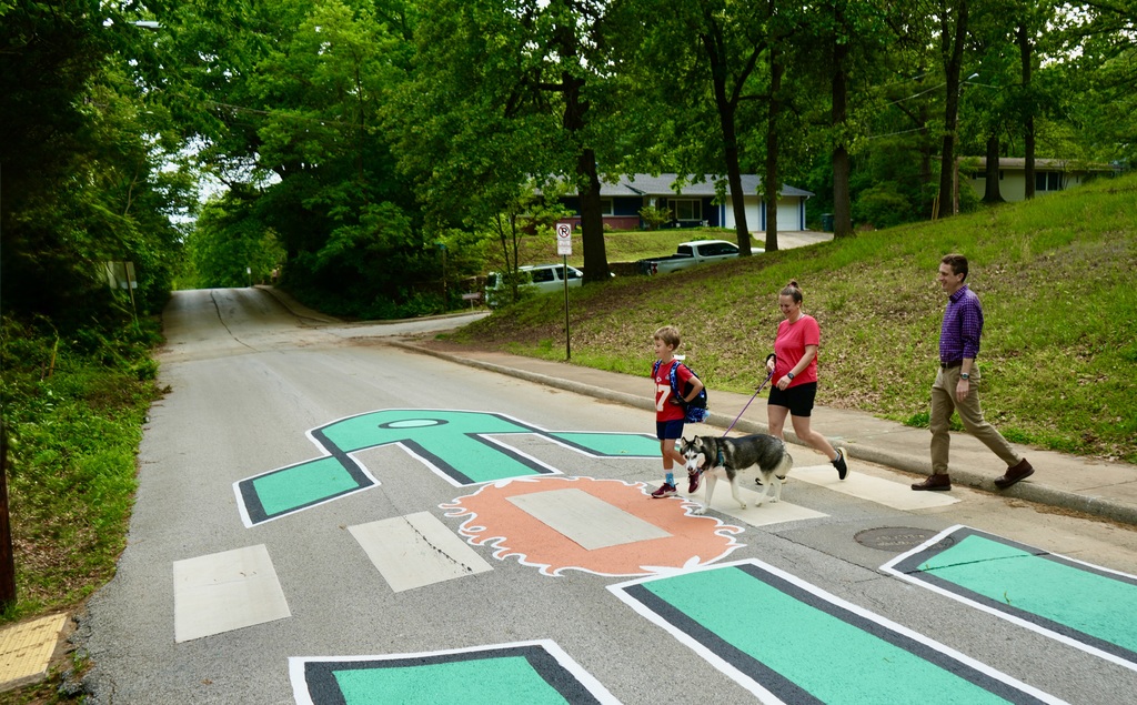 Three people, one with a dog, walk along a street with a green cross symbol painted on the road.