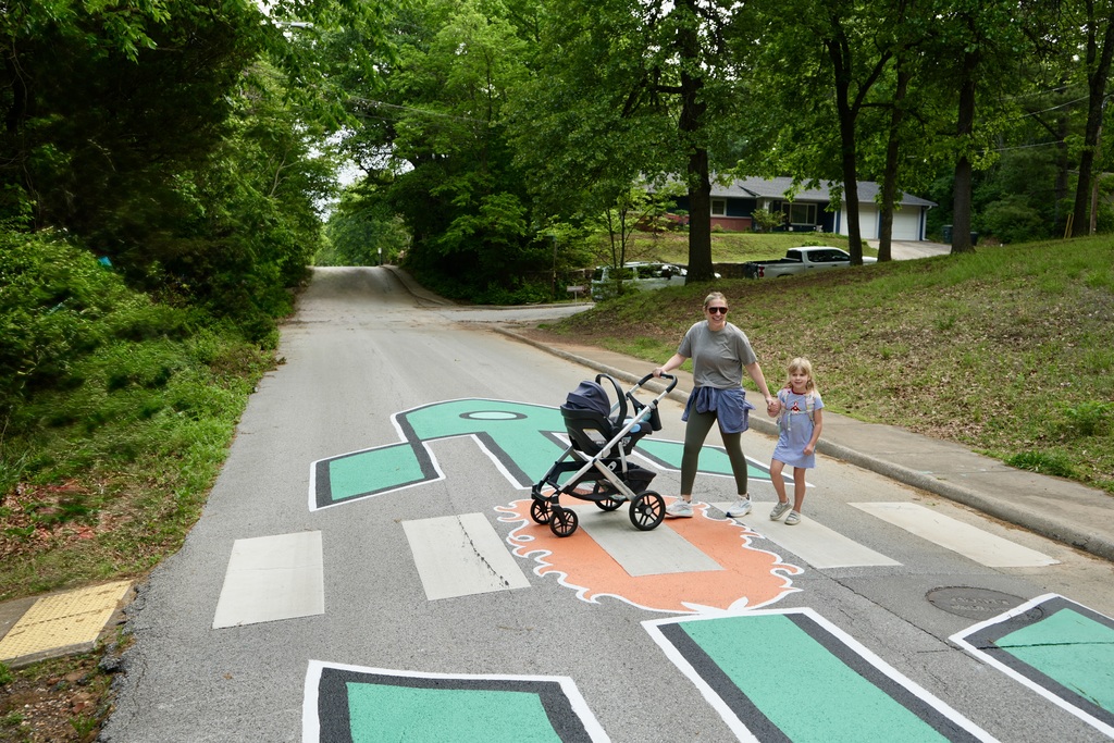A woman in a gray shirt pushes a stroller with a baby while walking on a pedestrian crossing.