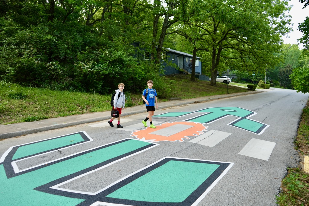 Two children are walking on the road with a painted design. Trees and bushes line the sides.