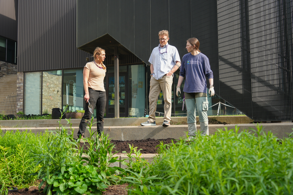 Three people, two women and a man, are standing in a garden with plants in front of a building.
