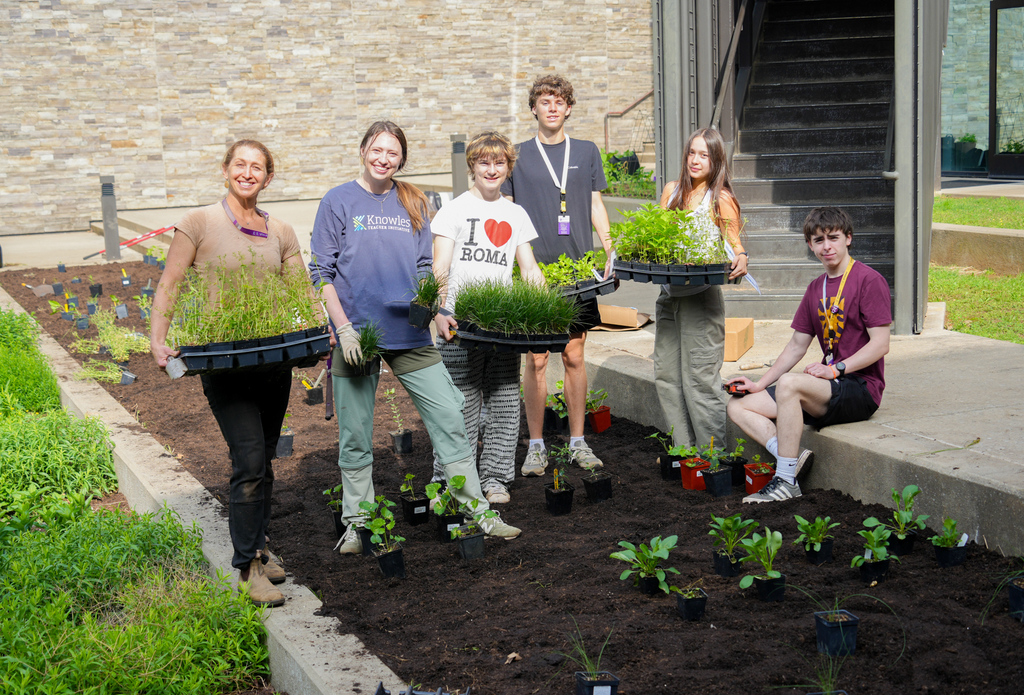 A group of people in a garden, each holding a tray of plants, with stairs and a brick wall in the background.