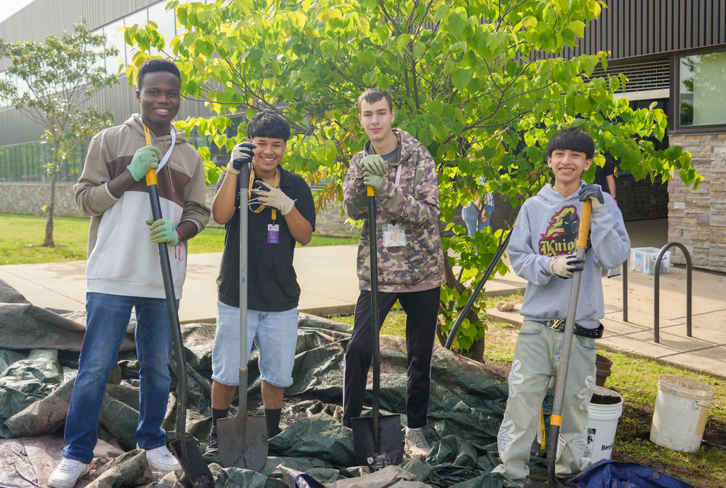 Four individuals, wearing gloves, standing around a tree. They are holding shovels.