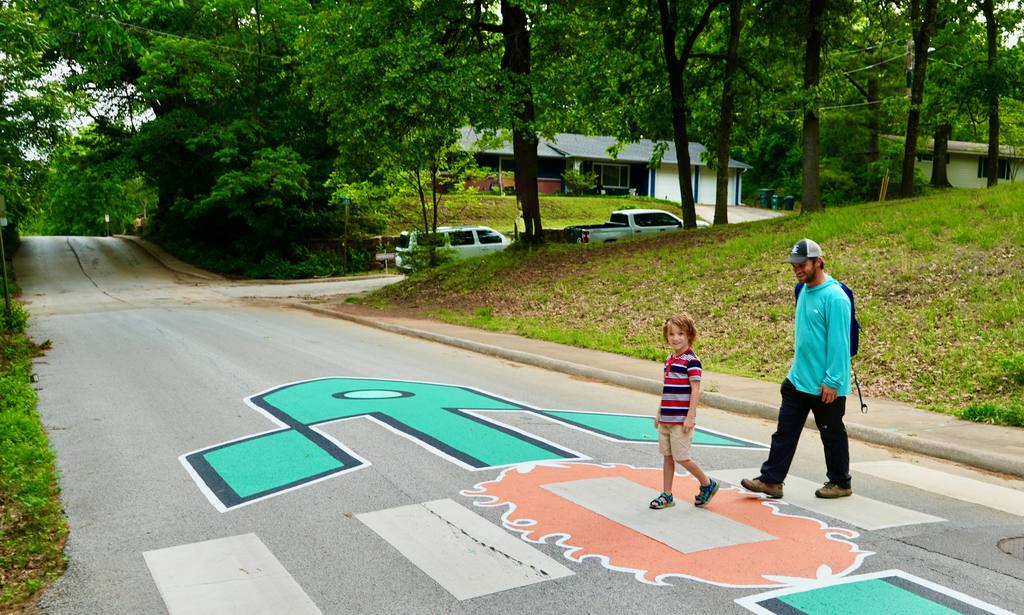 A man and a child cross a street with a crosswalk featuring large painted letters and numbers.