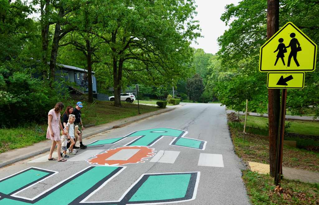 People crossing a road with a painted crosswalk and a pedestrian crossing sign. Trees and bushes on either side.