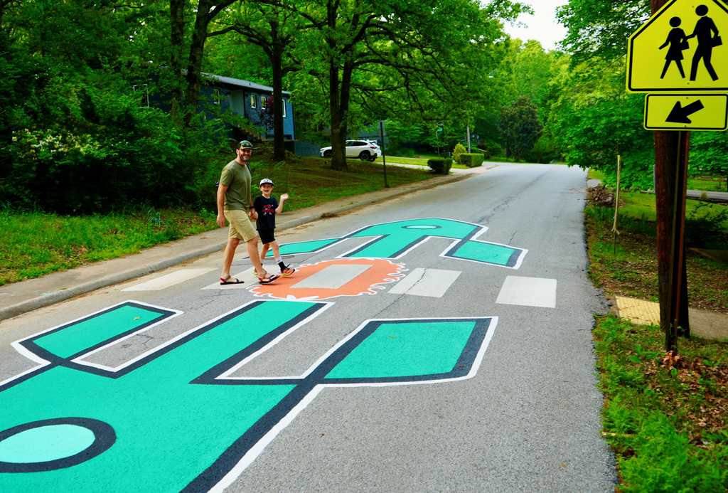 An adult and child cross a street with a painted pedestrian crossing. Trees and greenery line the road.