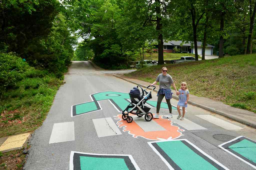 A parent pushes a stroller with a child down a road with painted letters and arrows. Trees line the sides.