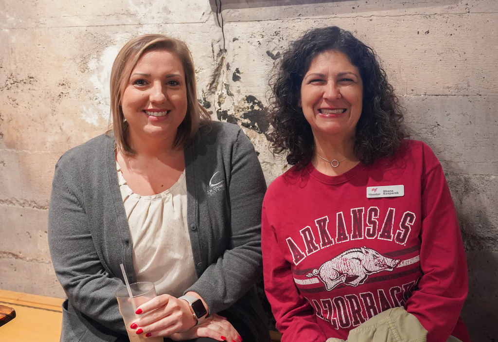 Two women sit together, smiling and holding drinks. One wears a red long-sleeve shirt, the other a gray cardigan.