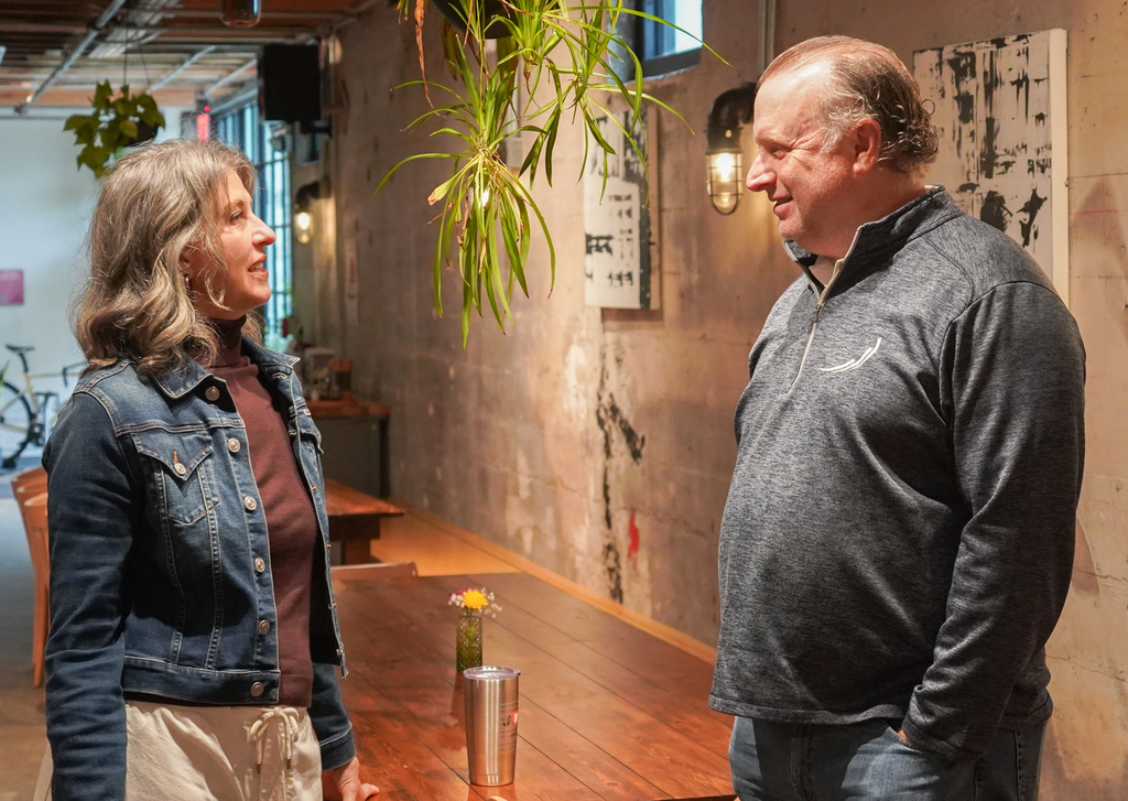 A man and a woman face each other in an indoor area with a wall-mounted artwork.