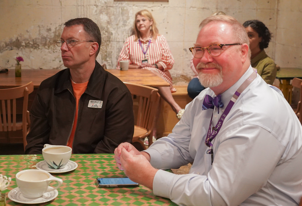 Three people sit at a table, one in a brown jacket, another in a white shirt with a bowtie.