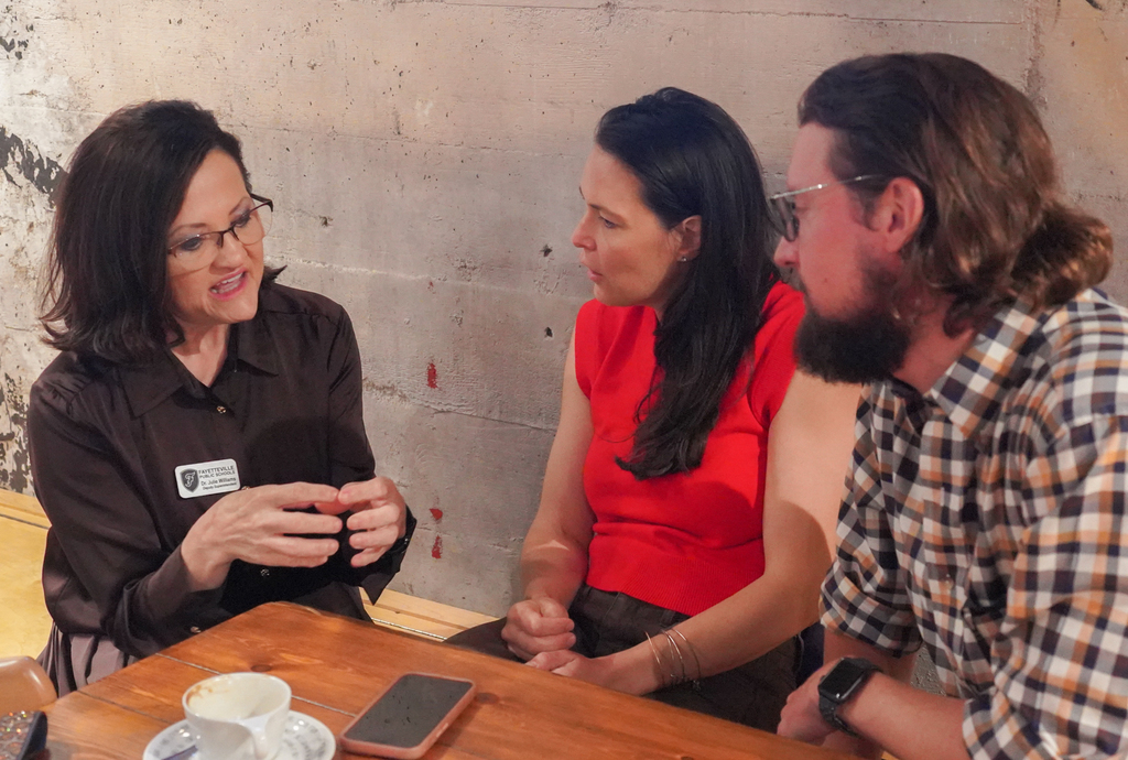Three people around a wooden table in a room, two women and one man. One woman wears glasses, another wears a red top.