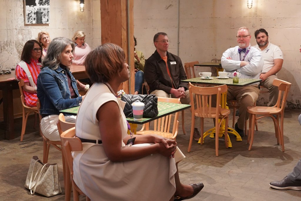 Group of people in a cafe, seated around tables. Some are conversing. One woman sits on a chair, facing the camera.