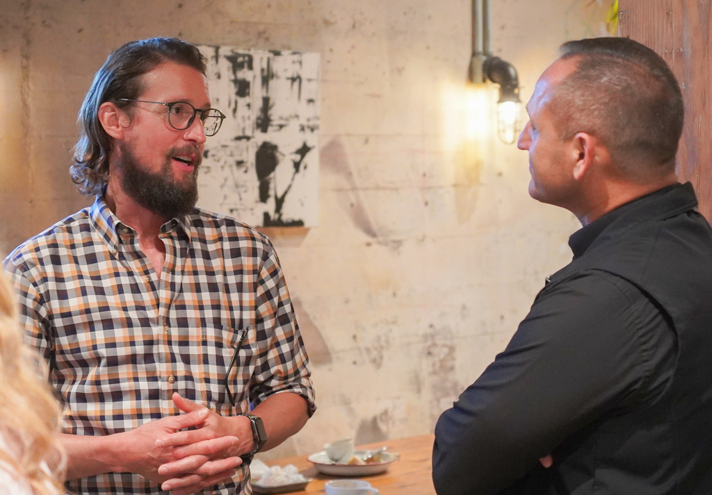 Two men face each other. One wears a checkered shirt and glasses. The other wears a black jacket.