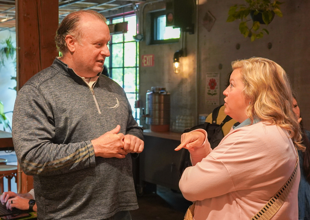 An adult man and woman speak animatedly in a room with a coffee bar, windows, and a light fixture.