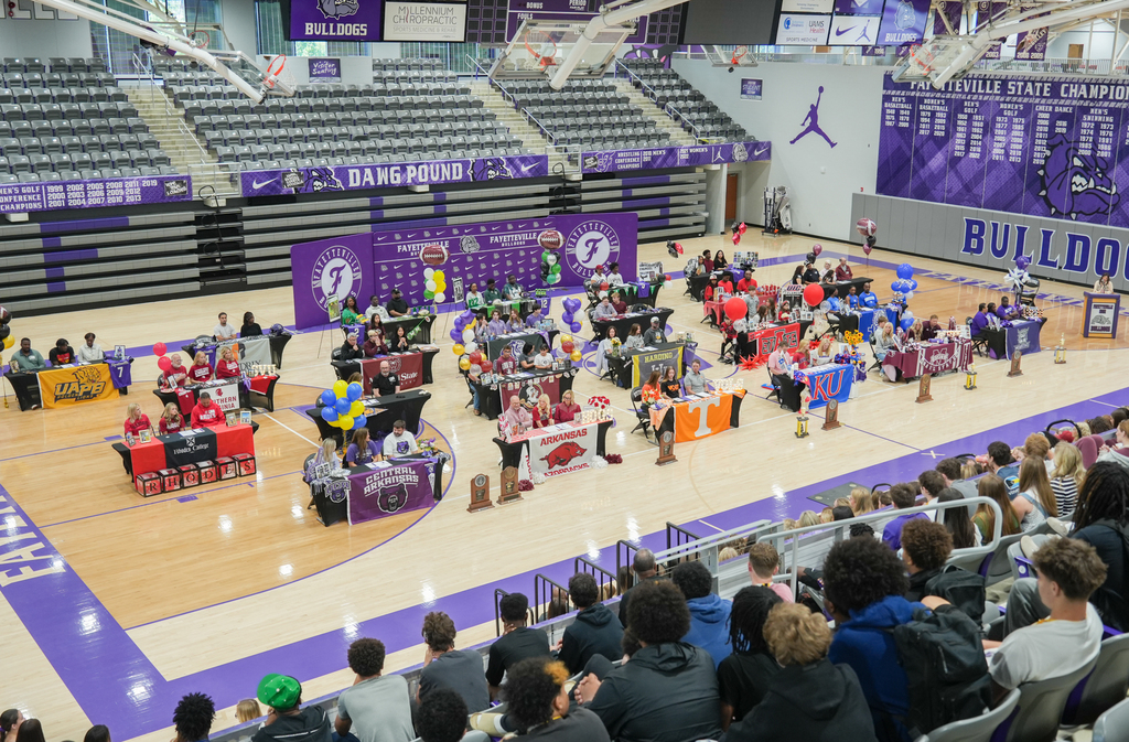 An indoor basketball court hosts a large event with numerous tables, balloons, and a crowd of people.
