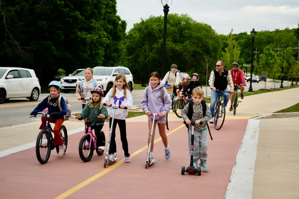 A group of kids ride bikes and scooters on a path in a park, with cars passing by.