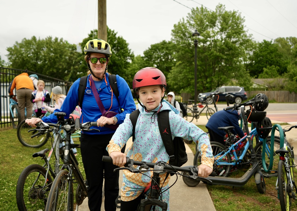 An adult and child stand on a sidewalk, both wearing helmets, with bicycles. Behind them, people are standing near parked bicycles. Trees and a fence are in the background.