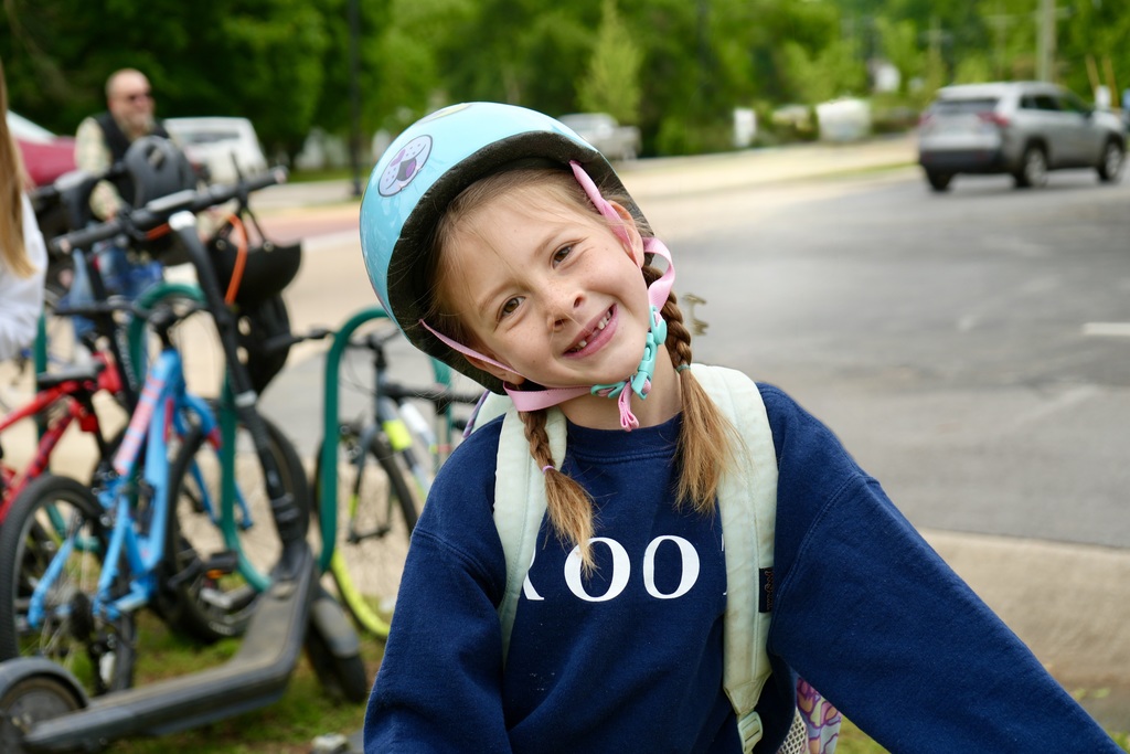 A young girl in a helmet smiles on the sidewalk, with bicycles behind her and a car on the road.