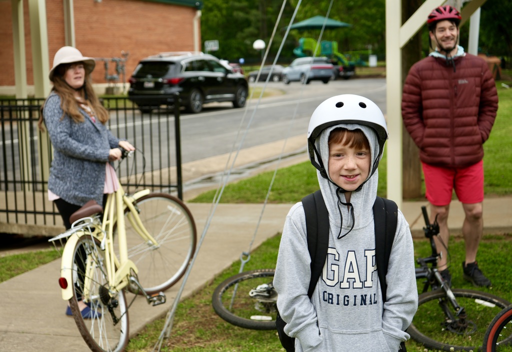 Three people stand on a sidewalk. A girl with a white hat holds a yellow bicycle. A boy in a gray hoodie stands in front of them. A man in a red coat stands next to a black car.
