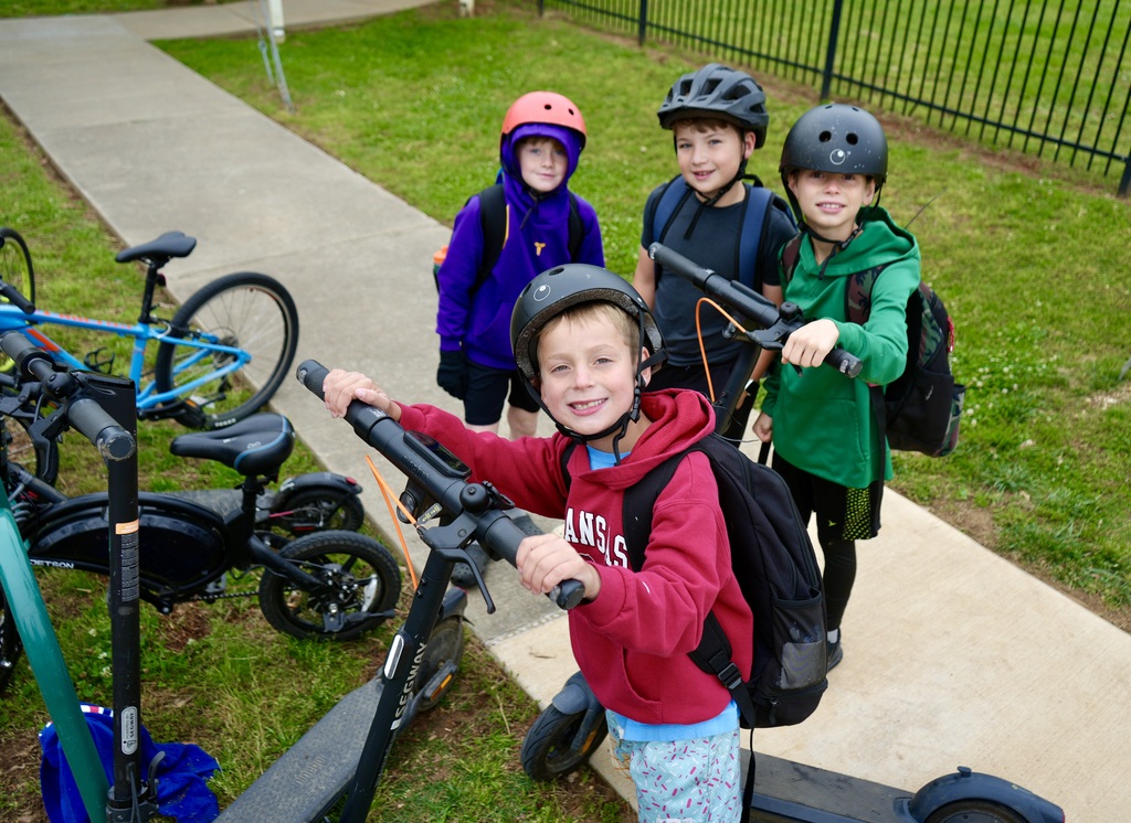 Four children, one wearing a helmet, stand on a sidewalk near e-scooters, with a grassy area and fence in the background.