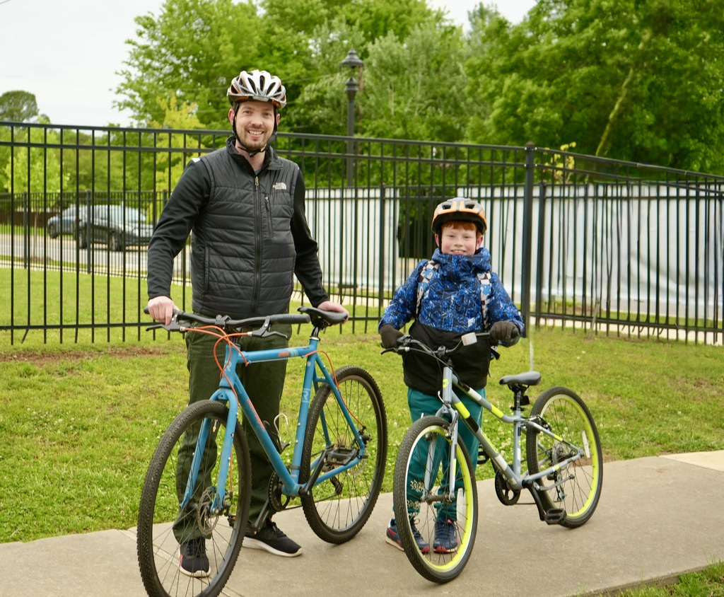 A man and a child standing on a sidewalk, both with helmets. The man holds a blue bike, and the child holds a green bike. Behind them is a black fence.