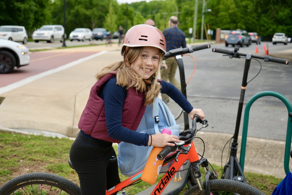 A child with a pink helmet and safety vest rides a red bicycle on grass near traffic.