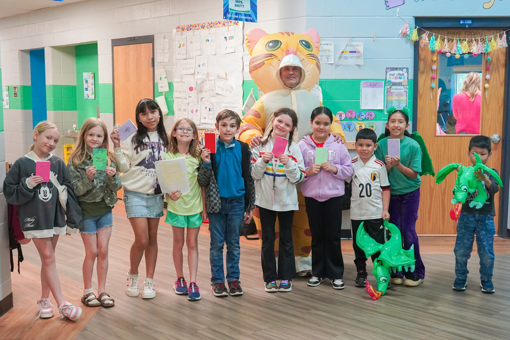 Kids stand together in a classroom with a mascot. They hold objects and wear casual clothes.
