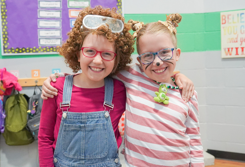 Two girls with glasses and matching accessories pose together in a classroom. One wears a pink shirt and overalls; the other wears a striped shirt.