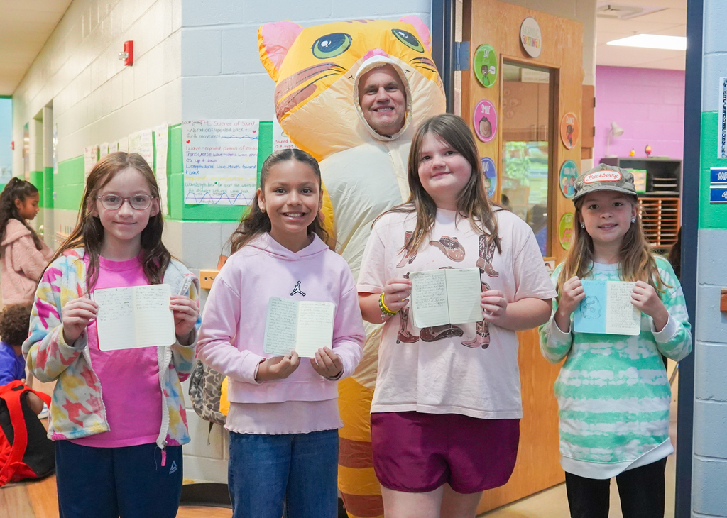 Five girls and a person dressed in a cat costume hold white papers in a classroom.
