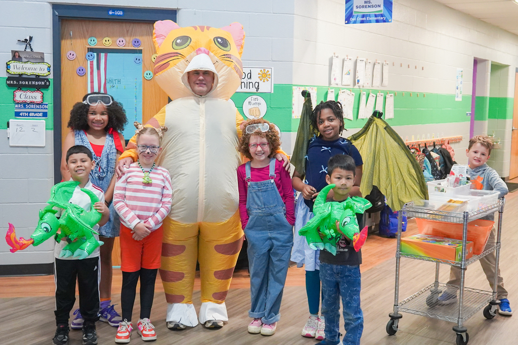 Several children stand with a mascot and one holds a toy dragon. Behind them, a door and classroom walls are visible.