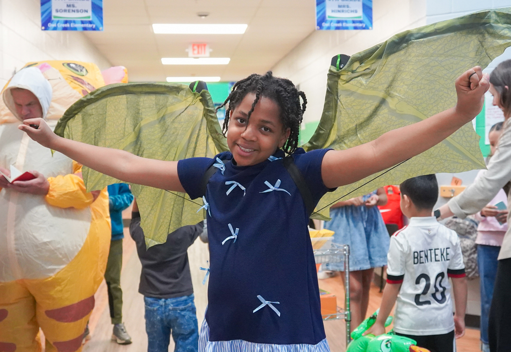 A young girl with wings attached to her back smiles and poses with arms outstretched.