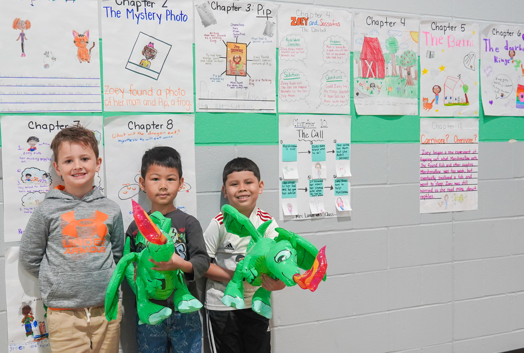 Three children stand together, each holding a green puppet. Behind them, a white wall displays various colorful posters.