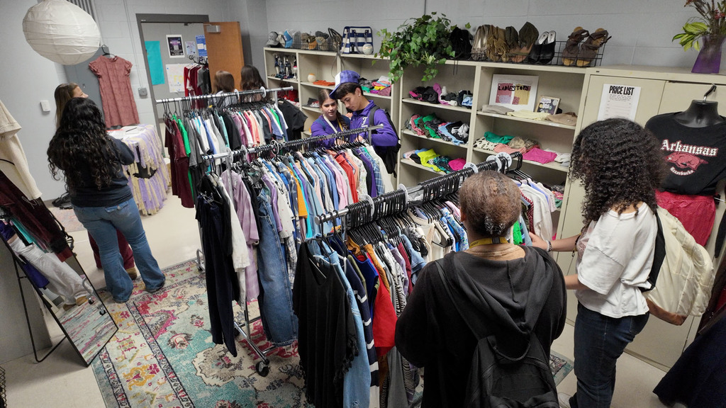 People browse a clothing store with racks of shirts, jeans, and jackets, with a rug on the floor.