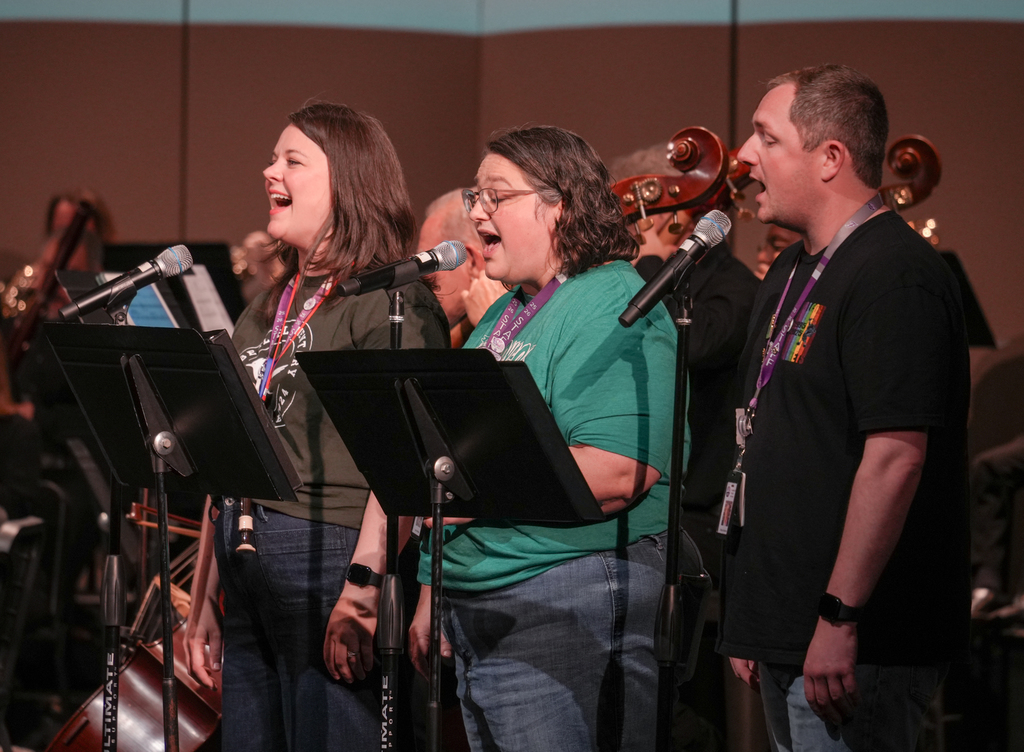 People stand in front of music stands with microphones during an event; three women sing and a man stands nearby.